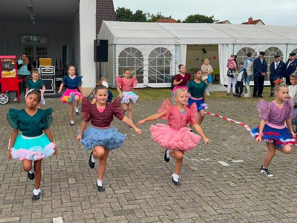 Gruppe von Kindern in bunten Tutu-Kleidern tanzt synchron auf einem gepflasterten Platz vor weißen Festzelten.