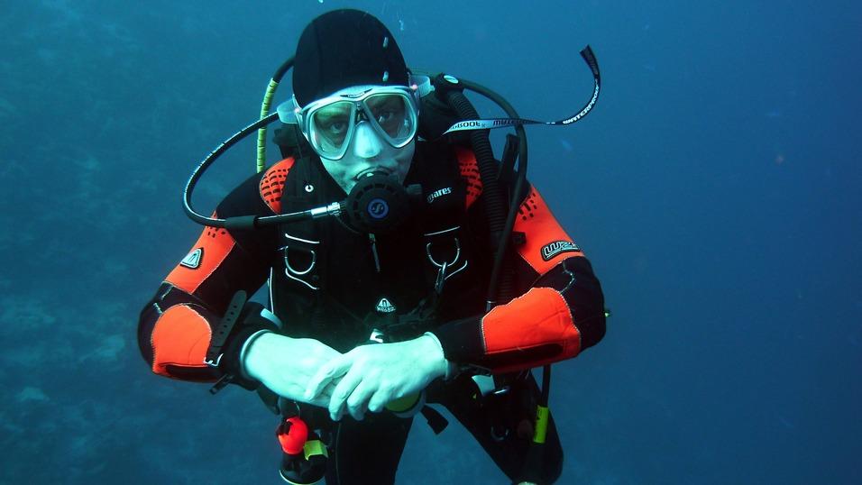 Taucher in einem schwarzen Neoprenanzug mit roten Akzenten schwebt unter Wasser und blickt direkt in die Kamera.