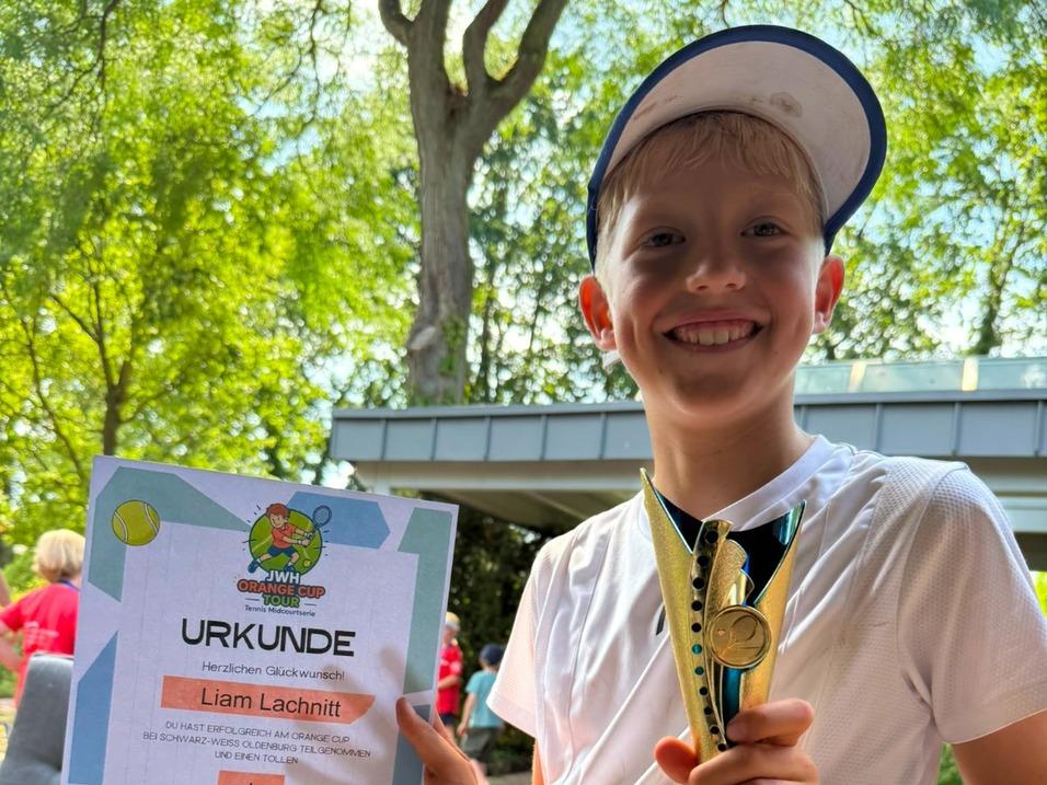 Junge mit weißer Schirmmütze hält eine Urkunde und einen Pokal in der Hand und lächelt in die Kamera.