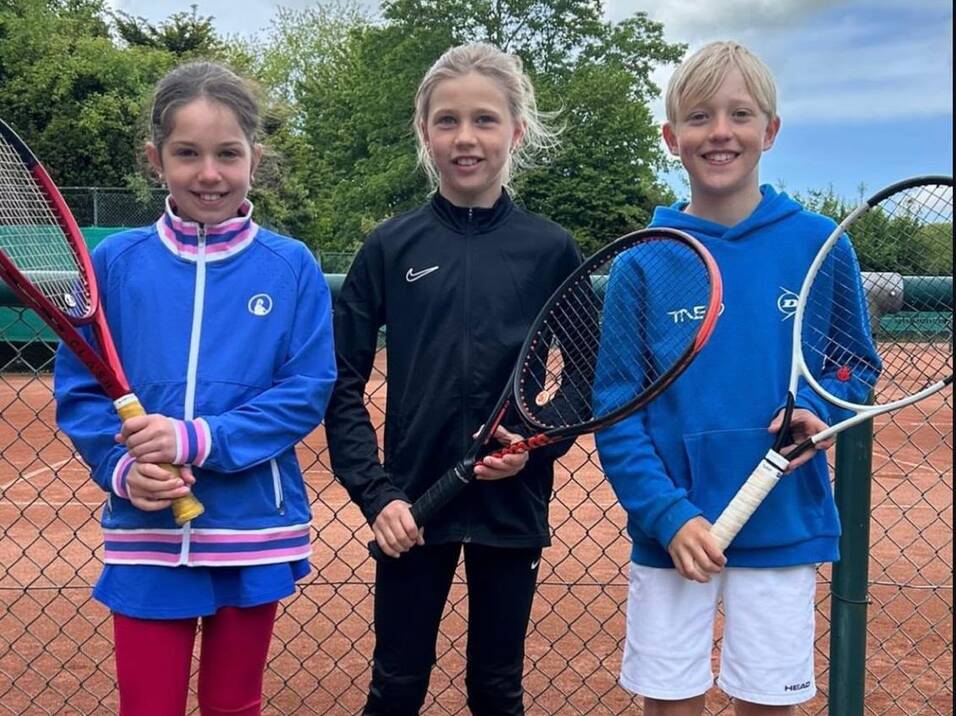 Drei Kinder mit Tennisschlägern stehen nebeneinander auf einem Tennisplatz vor einem Zaun.