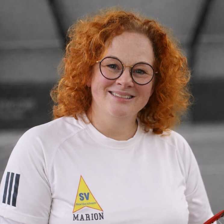 Portr&auml;t einer l&auml;chelnden Frau mit r&ouml;tlichen Haaren, Brille und wei&szlig;em Oberteil, vor einem unscharfen Hintergrund.