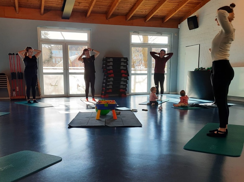 Gruppe von Frauen mit Kindern während einer Fitnessstunde in einem Raum mit Übungsmatten und Fenstern.
