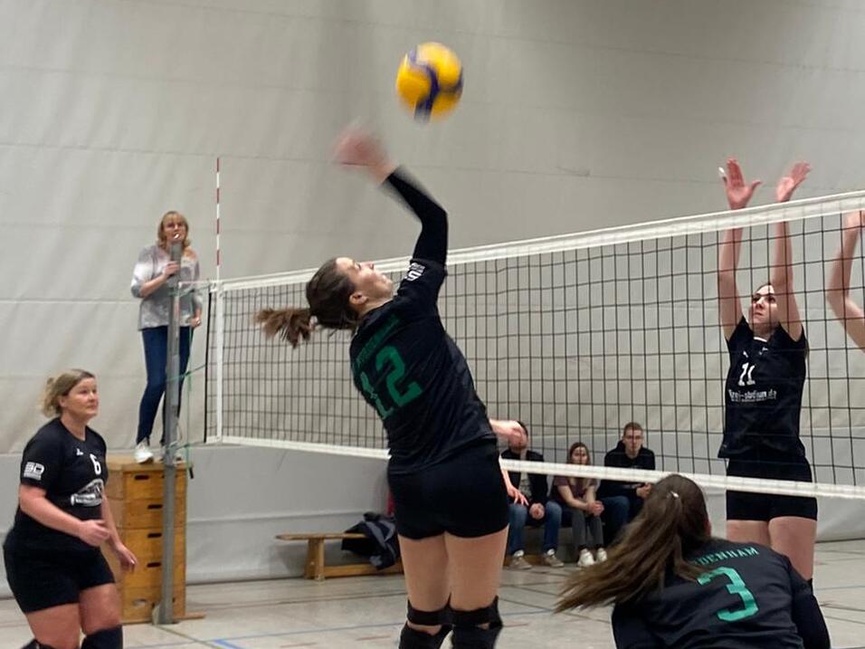 Spielszene im Volleyball mit einer Spielerin, die zum Sprung ansetzt, während drei Gegnerinnen blocken. Zuschauer im Hintergrund.