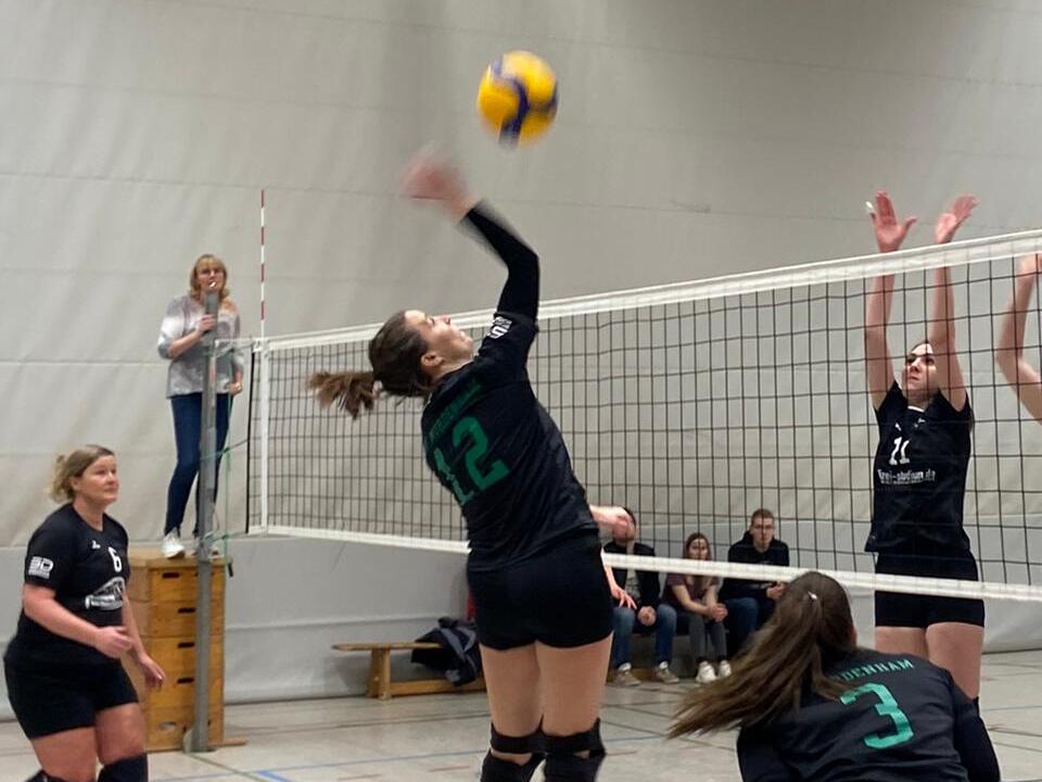 Spielszene im Volleyball mit einer Spielerin, die zum Sprung ansetzt, während drei Gegnerinnen blocken. Zuschauer im Hintergrund.