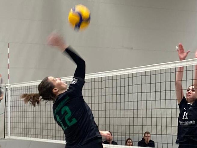 Spielszene im Volleyball mit einer Spielerin, die zum Sprung ansetzt, w&auml;hrend drei Gegnerinnen blocken. Zuschauer im Hintergrund.