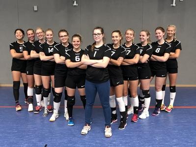 Mannschaftsbild von zw&ouml;lf weiblichen Volleyballspielerinnen in schwarzen Trikots auf einem Sportboden in einer Halle.