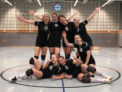 Gruppensfoto von sechs Volleyballspielerinnen in schwarzen Trikots, fröhlich posierend in einer Sporthalle.