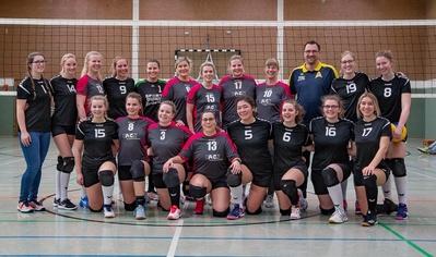 Gruppenfoto einer Volleyballmannschaft mit 15 Spielerinnen und einem Trainer, in Sportbekleidung auf einem Volleyballfeld.