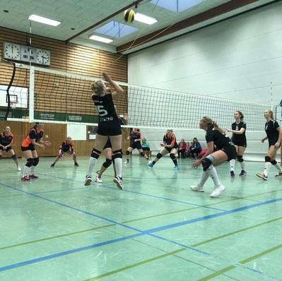 Volleyballspiel in der Halle: Spielerinnen in Schwarz und Orange, eine springt zum Aufschlag, andere bereit in Positionsspiel.