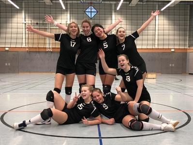 Gruppensfoto von sechs Volleyballspielerinnen in schwarzen Trikots, fr&ouml;hlich posierend in einer Sporthalle.