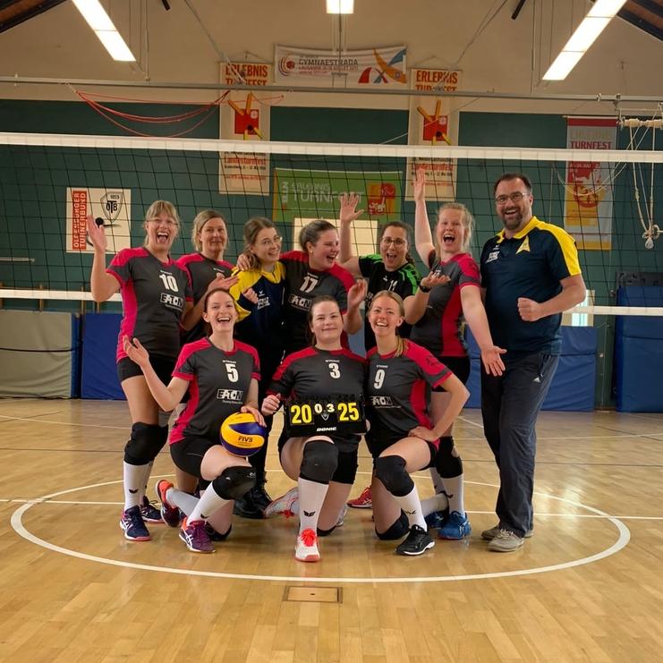 Gruppenselfie eines Volleyballteams auf dem Spielfeld, lächelnd mit einem Scoreboard, das 20:25 zeigt.