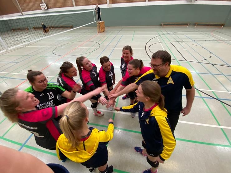 Gruppenkreis von Volleyballspielerinnen und Trainer vor einem Spiel in einer Sporthalle, Hände zum Vereinsgruß aufgestreckt.