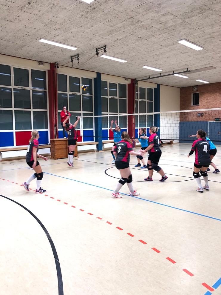 Volleyballtraining mit mehreren Spielerinnen in der Sporthalle, eine Spielerin springt für einen Angriff.