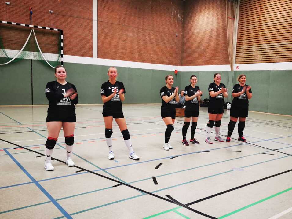 Sechs Frauen in schwarzen Trikots klatschen in einer Sporthalle, mit einem Handballtor im Hintergrund.