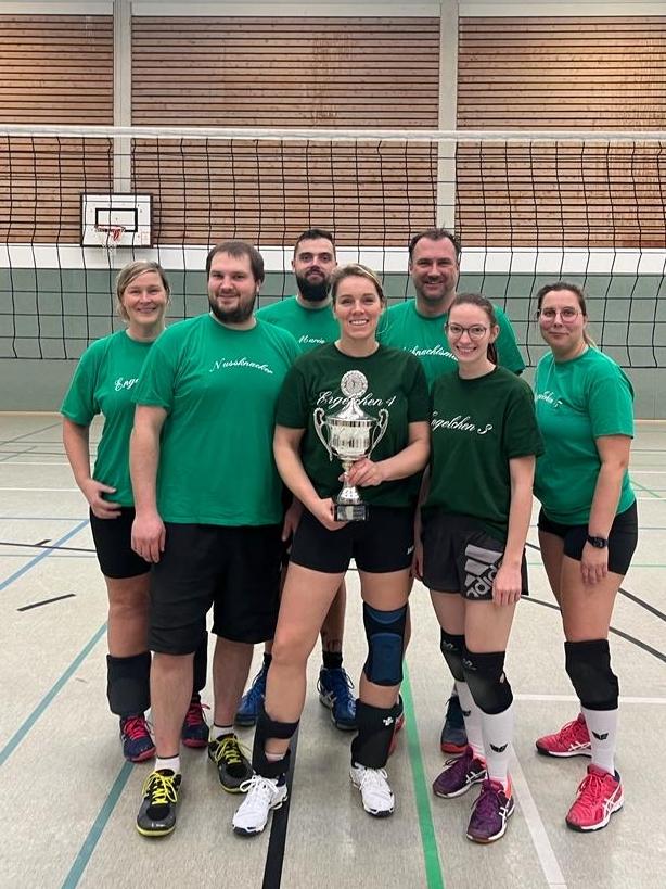 Gruppenfoto von sieben Volleyballspielern in grünen T-Shirts, die einen Pokal in der Sporthalle präsentieren.