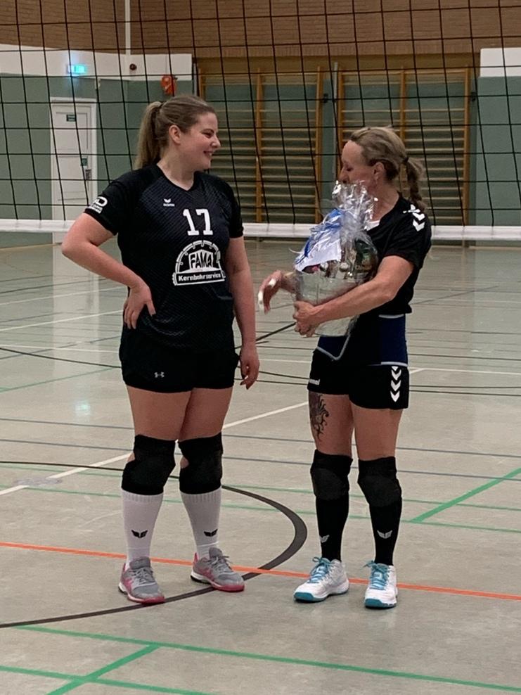 Zwei Volleyballspielerinnen unterhalten sich in der Halle, eine hält einen mit Geschenkverpackung dekorierten Preis.