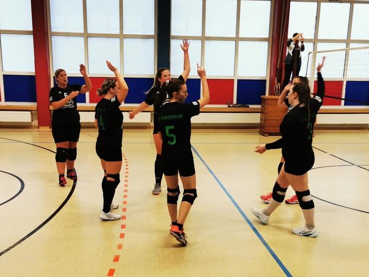 Sechs Volleyballspielerinnen in schwarzen Trikots feiern in einer Sporthalle nach einem Punktgewinn.