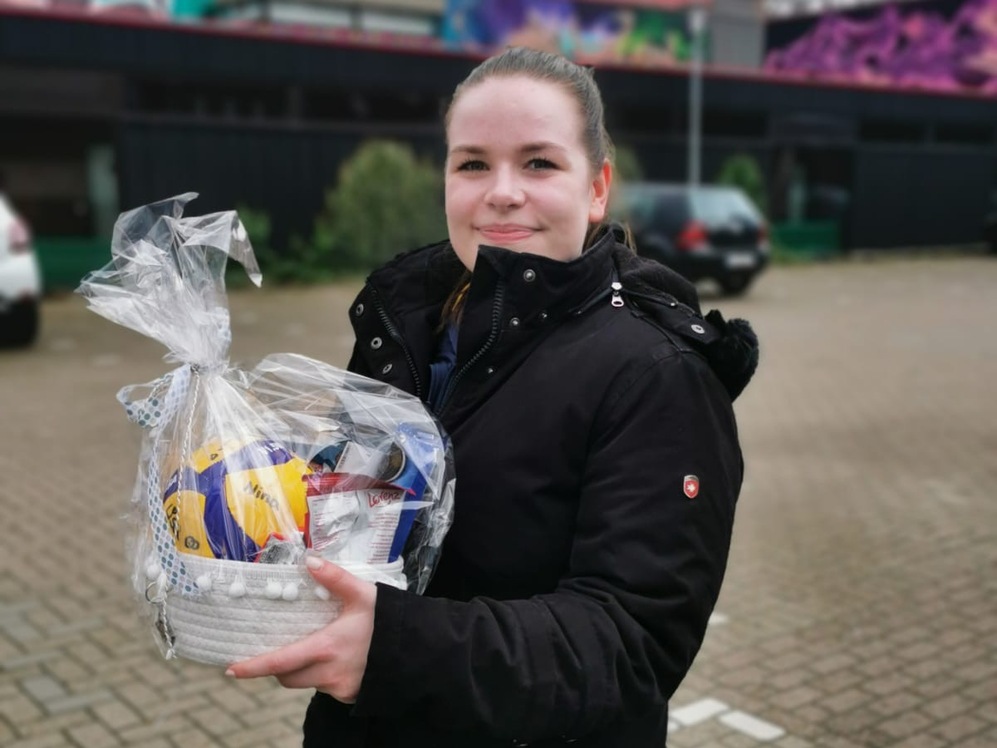 Frau mit schwarzer Jacke h&auml;lt einen Geschenkkorb in der Hand, stehend auf einem Parkplatz mit grauer Pflasterung.