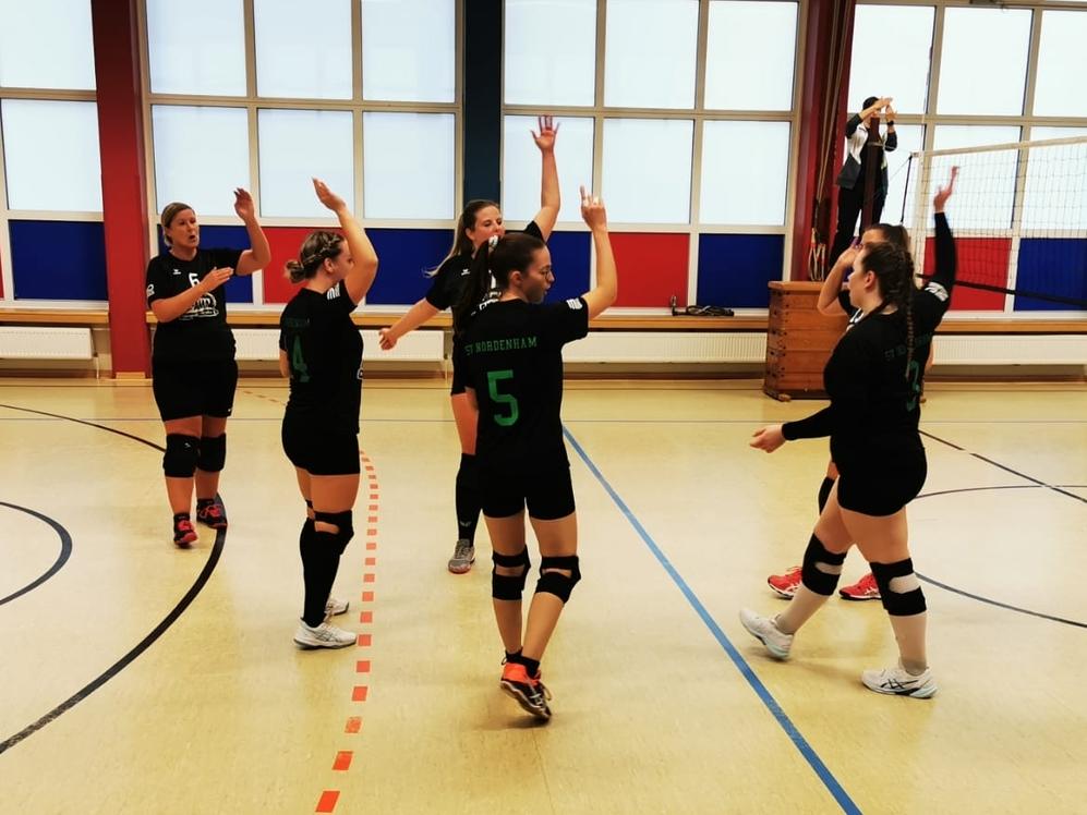 Sechs Volleyballspielerinnen in schwarzen Trikots feiern in einer Sporthalle nach einem Punktgewinn.