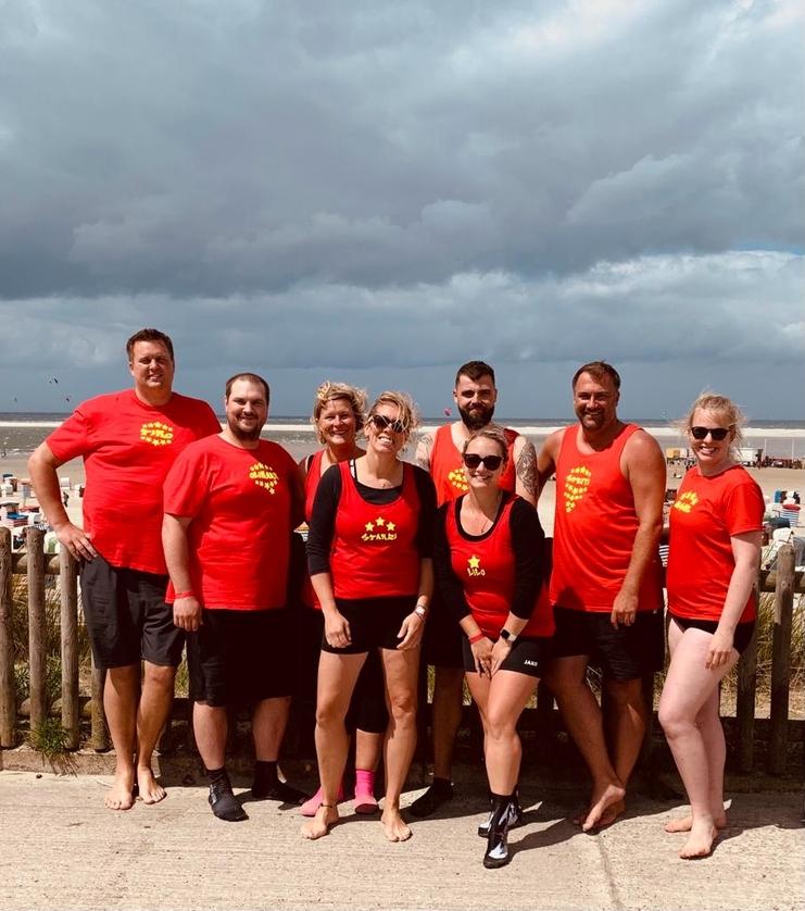 Gruppe von acht Personen in roten T-Shirts am Strand, mit Wolken und Meer im Hintergrund.