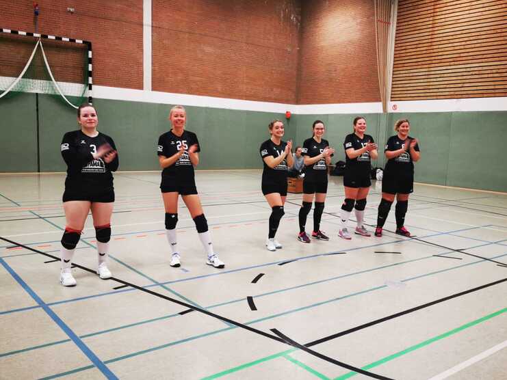 Sechs Frauen in schwarzen Trikots klatschen in einer Sporthalle, mit einem Handballtor im Hintergrund.