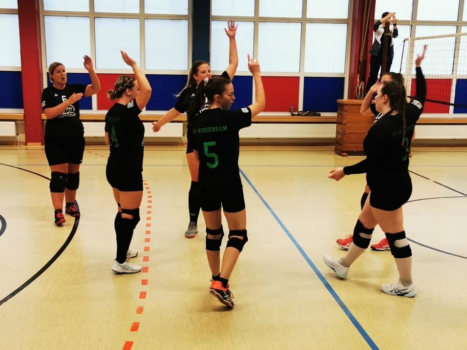 Sechs Volleyballspielerinnen in schwarzen Trikots feiern in einer Sporthalle nach einem Punktgewinn.
