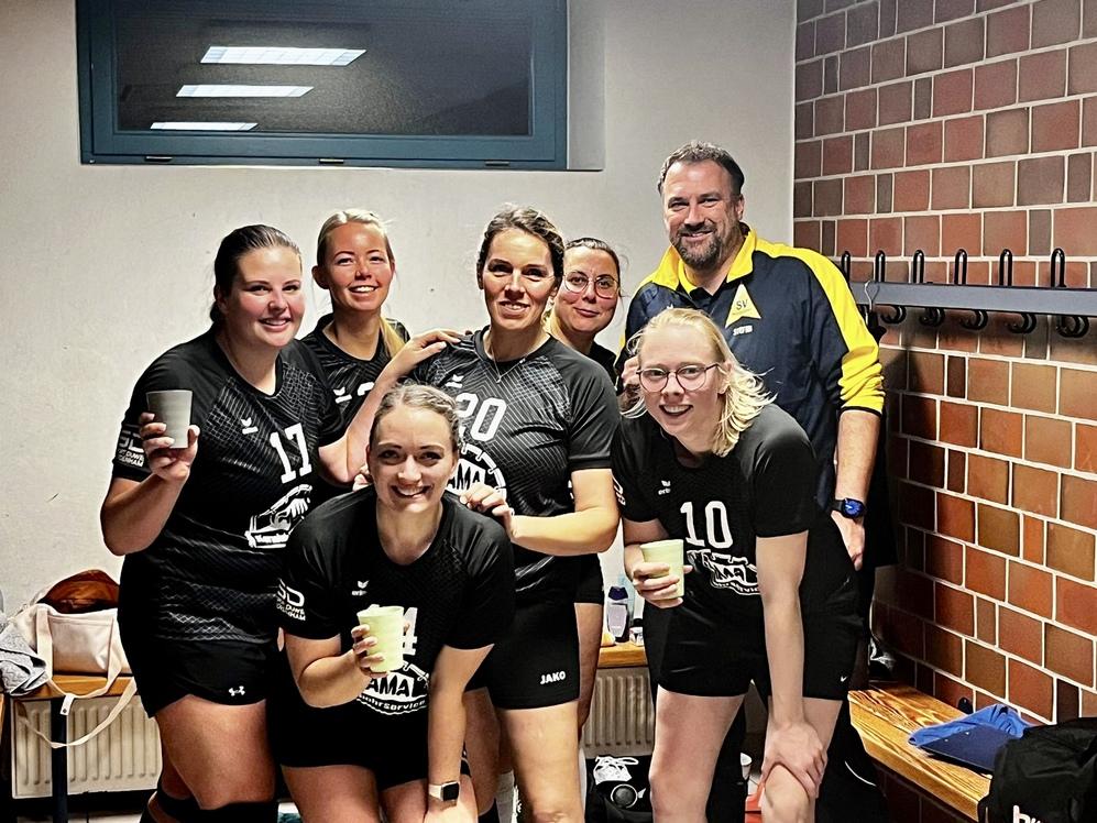 Sechs weibliche Sportler in schwarzen Trikots und einem Trainer l&auml;cheln in einer Umkleide mit Bechern in der Hand.