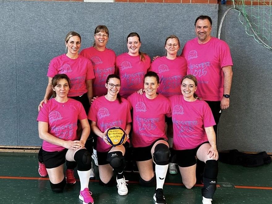 Gruppenfoto von acht Volleyballspielern in pinken Trikots mit einem Ball in der Mitte, vor einer Sportwand.