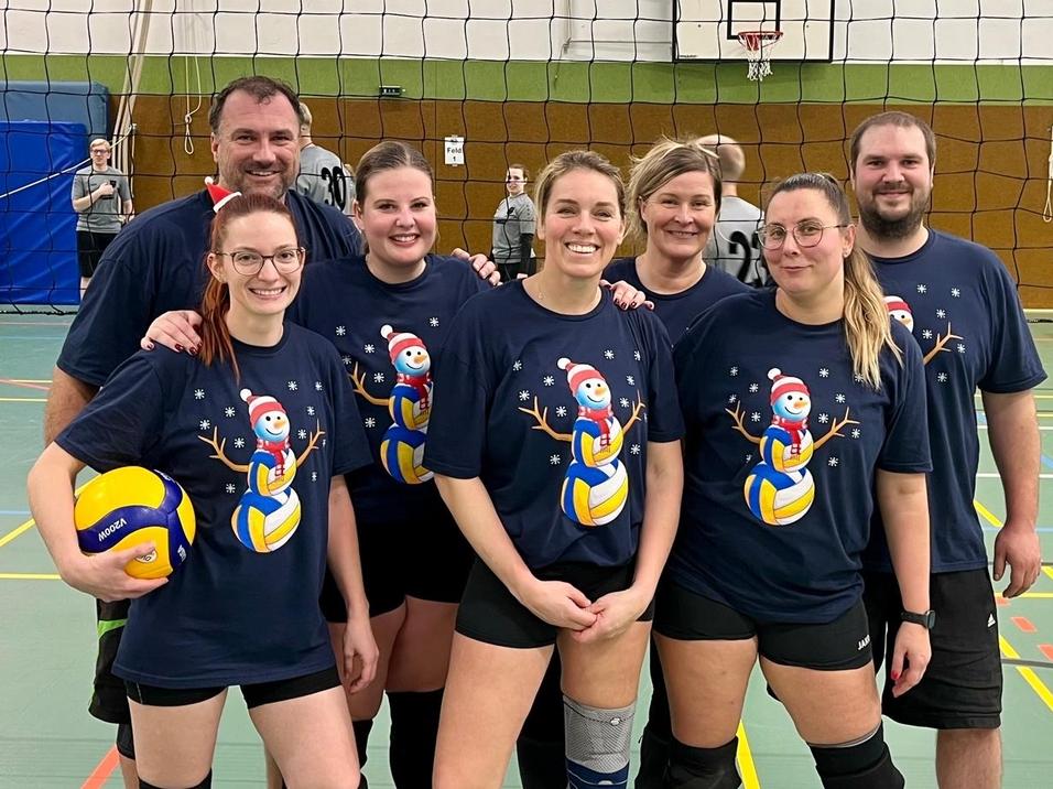 Gruppe von sieben Volleyballspielern in weihnachtlichen T-Shirts vor einem Volleyballfeld, mit einem Ball in der Hand.