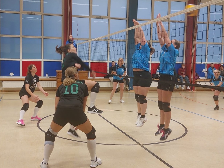 Volleyballspiel in der Halle: Spielerinnen springen gegen den Ball am Netz, mehrere Spielerinnen in Sportkleidung sind aktiv.