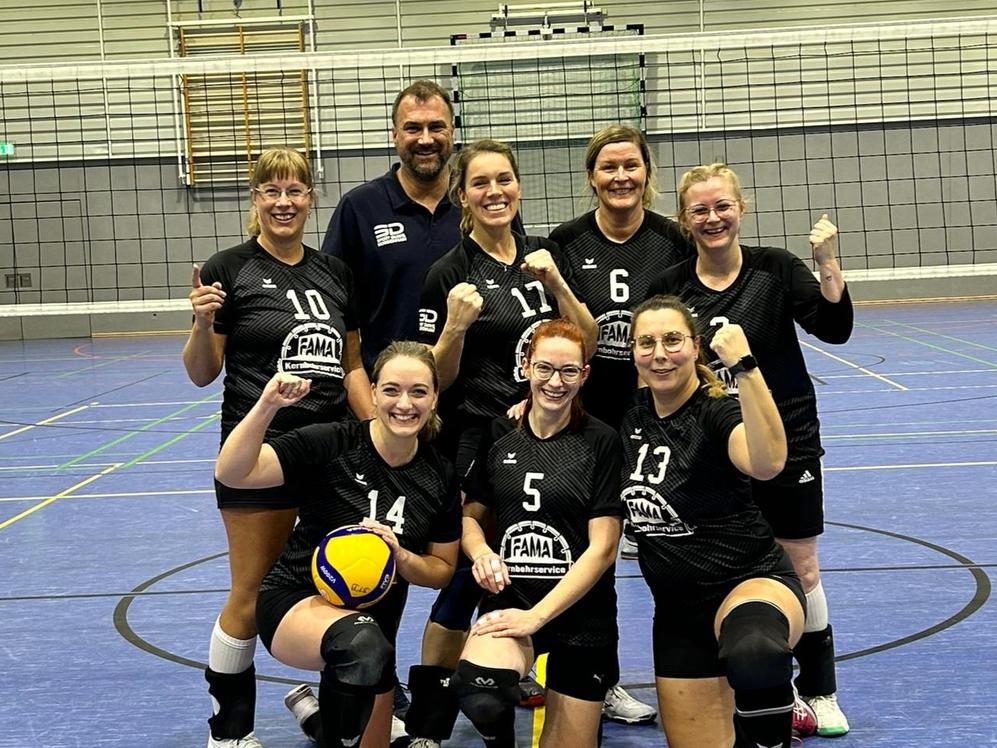Gruppe von sieben Volleyballspielerinnen in schwarzen Trikots posiert mit einem Volleyball und Punktestand auf dem Feld.
