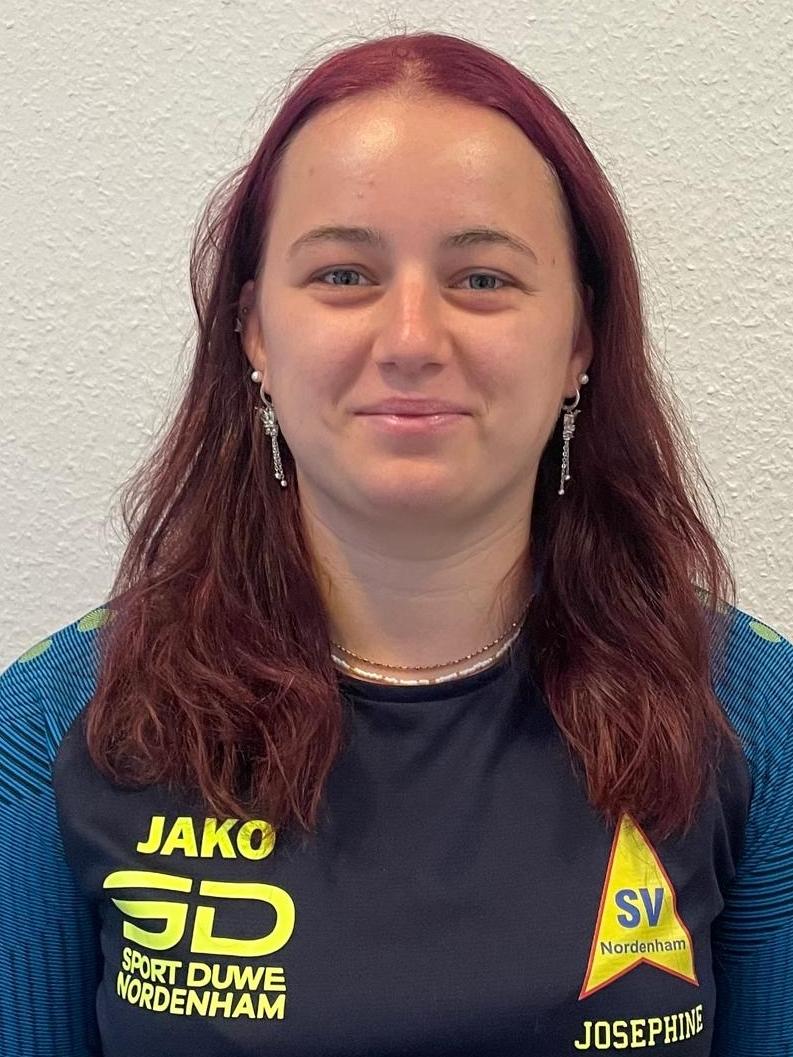 Porträt einer lächelnden Frau mit langen, braunen Haaren, gekleidet in ein sportliches Shirt vor einer weißen Wand.