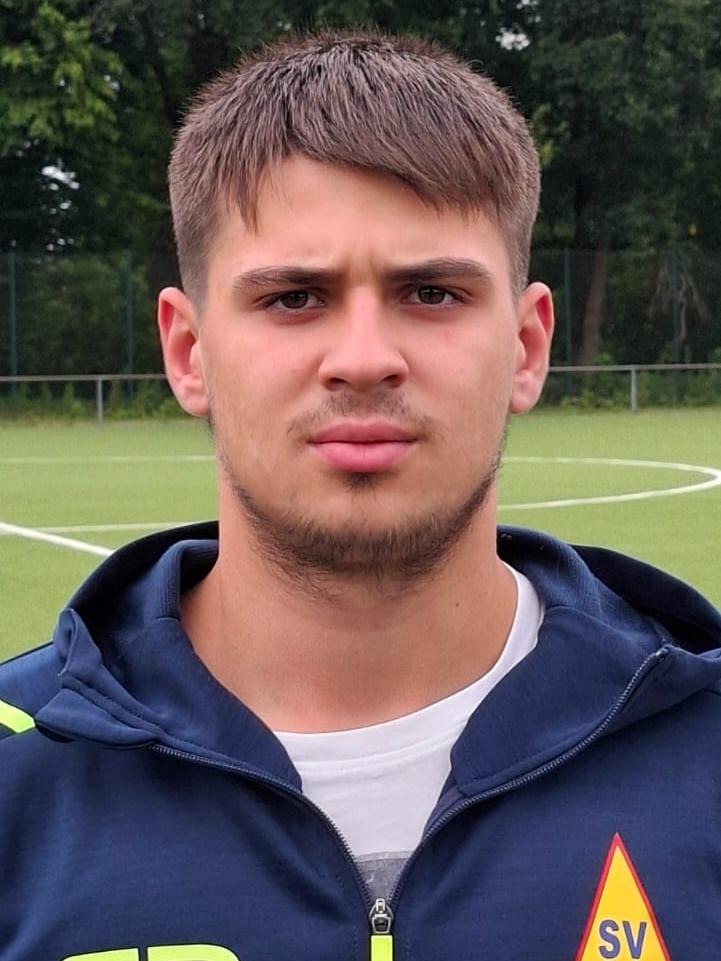 Junger Mann mit Kurzhaarschnitt, trägt einen blauen Trainingsanzug mit Vereinslogo, steht auf einem Fußballplatz.