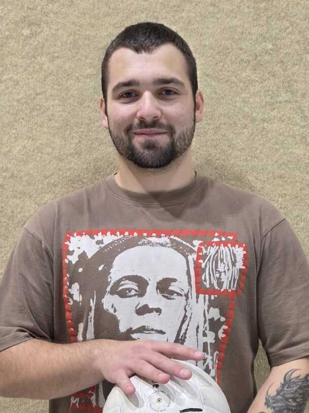 Junger Mann mit Kurzhaarschnitt, trägt ein T-Shirt mit Aufdruck, hält einen Ball in beiden Händen.