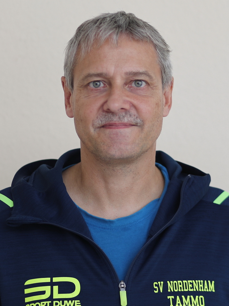 Mann in blauer Jacke mit gelben Akzenten, lächelt vor hellbeigem Hintergrund.