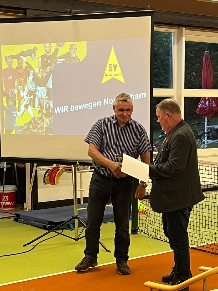 Zwei M&auml;nner &uuml;berreichen sich ein Dokument vor einer Projektionsleinwand mit dem Logo des SV Nordenham.