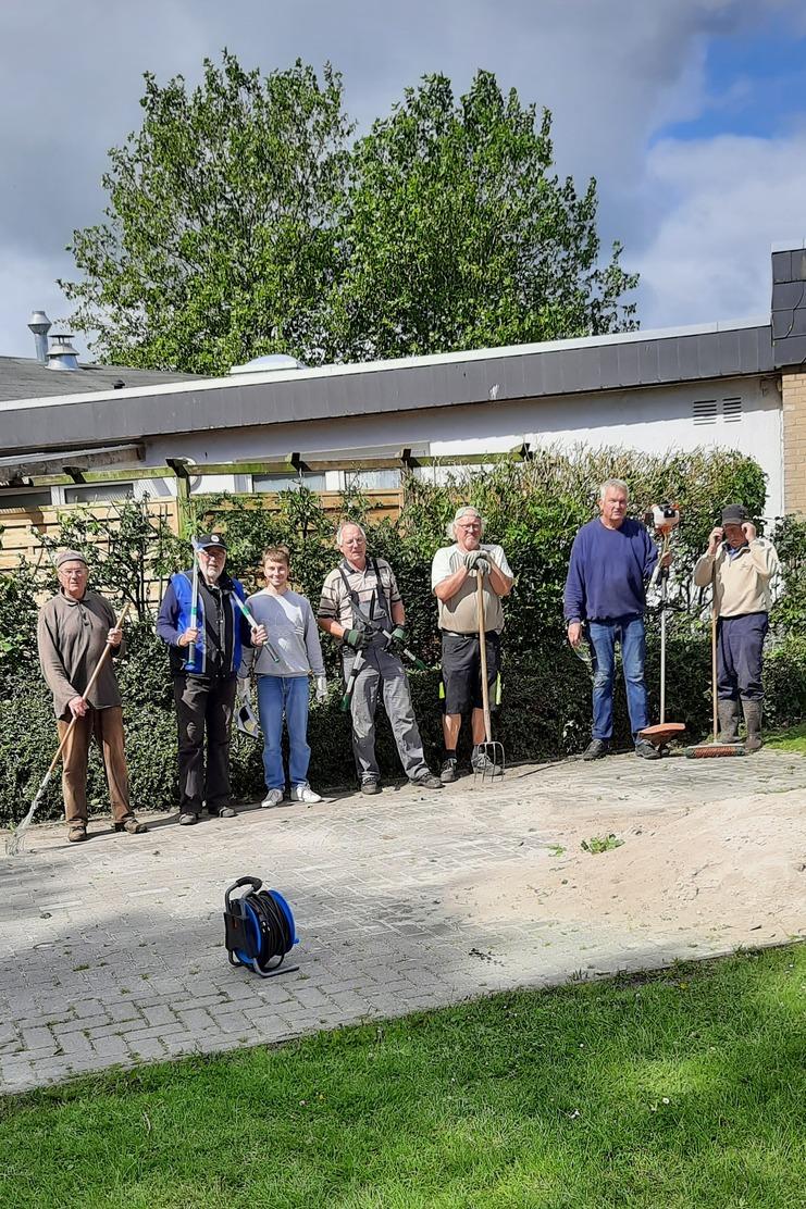 Gruppe von sieben Personen mit Gartenwerkzeugen steht entlang einer Hecke auf einem gepflasterten Weg im Freien.