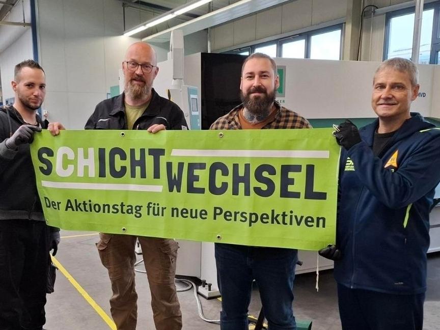 Vier M&auml;nner halten ein Banner mit der Aufschrift &bdquo;SCHICHTWECHSEL &ndash; Der Aktionstag f&uuml;r neue Perspektiven&ldquo; in einer Halle.
