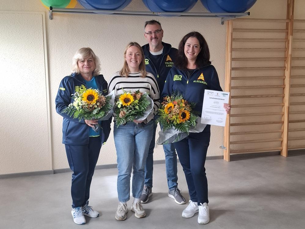 Drei Personen stehen in einem Raum und halten Blumen, w&auml;hrend sie fr&ouml;hlich in die Kamera blicken.