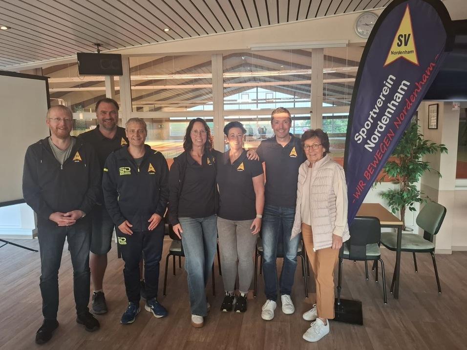 Gruppe von sechs Personen in Sportjacken vor einem Banner des SV Nordenham in einem Vereinsraum.