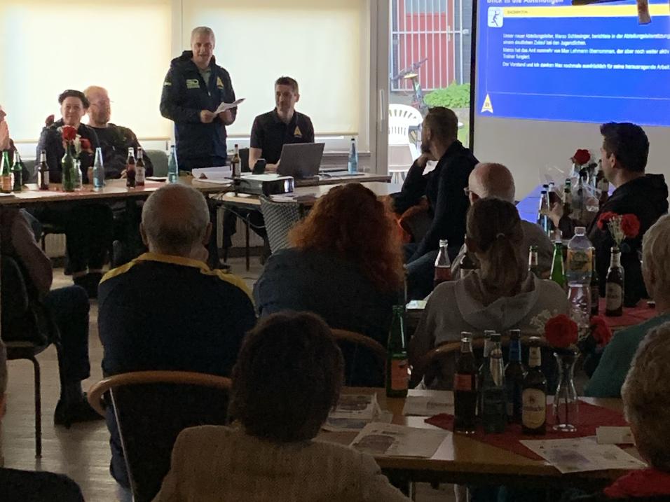 Person steht vor einem Bildschirm mit Präsentation in einem Raum mit mehreren sitzenden Zuhörern.