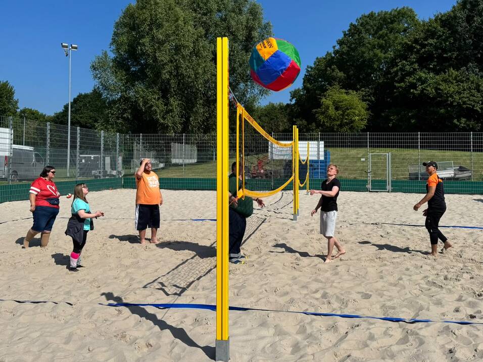 Mehrere Personen spielen auf einem Sandfeld mit einem gro&szlig;en bunten Ball &uuml;ber ein Netz.