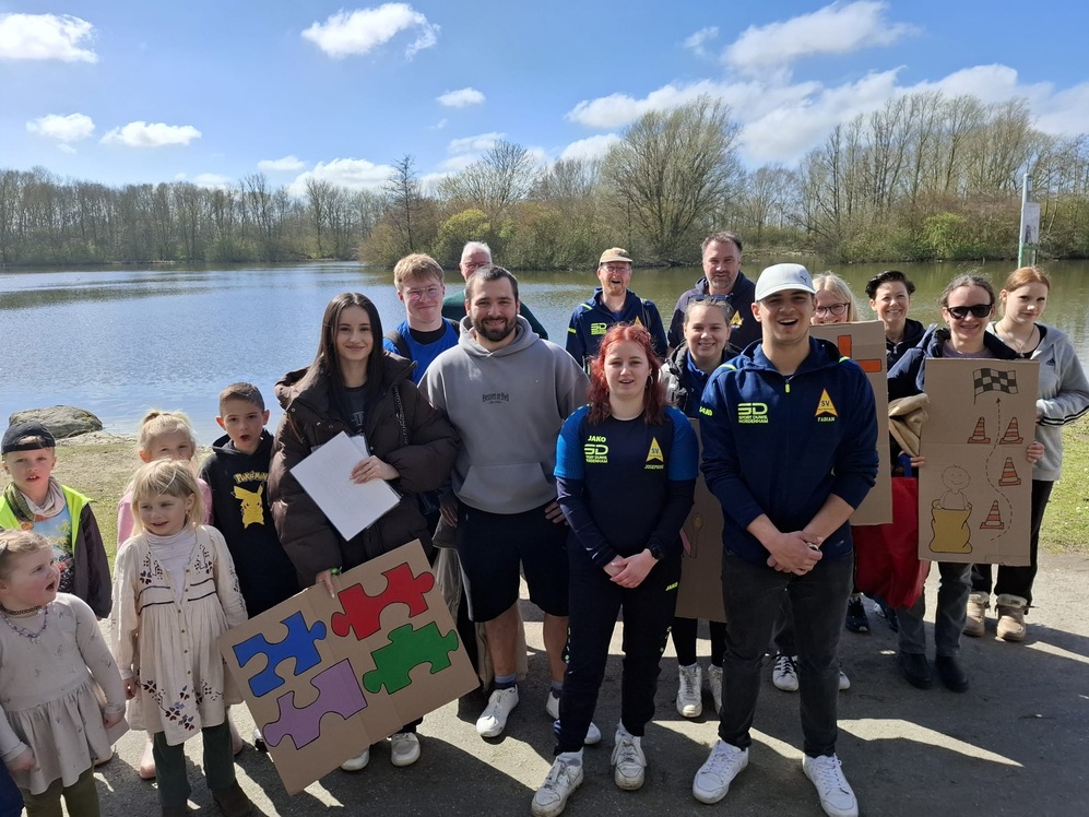 Vor einem See posiert eine Gruppe von Personen mit verschiedenen Puzzlest&uuml;cken in der Hand.