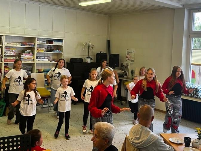 Kinder tanzen vor einer Gruppe &auml;lterer Menschen in einem Gemeinschaftsraum mit B&uuml;cherregal und gro&szlig;en Fenstern.