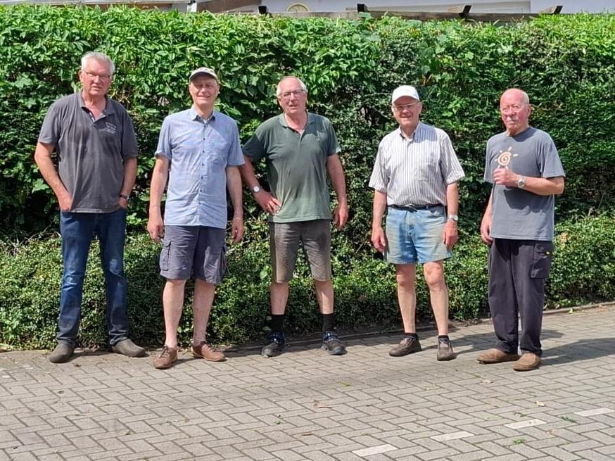 Fünf Männer stehen auf einem gepflasterten Platz vor einer hohen Hecke und einem flachen Gebäude.