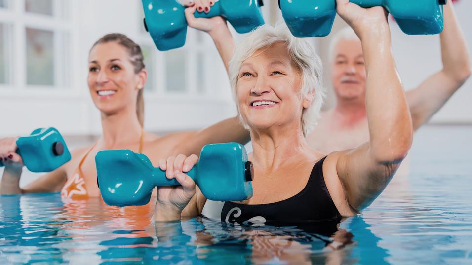 Gruppe von drei Personen im Wasser mit blauen Hanteln, l&auml;chelnd und aktiv bei einem Wasserfitnesskurs.