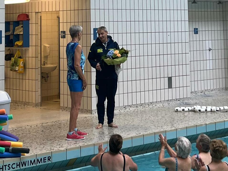 Zwei Personen am Beckenrand eines Schwimmbads, eine mit Blumenstrauß, umgeben von applaudierenden Schwimmern.