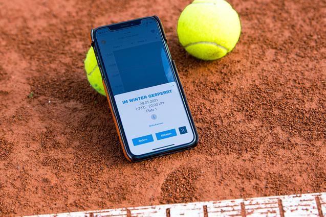 Smartphone mit Buchungsinfo liegt auf einem Tennisplatz, neben zwei gelben Tennisbällen auf rotem Sand.