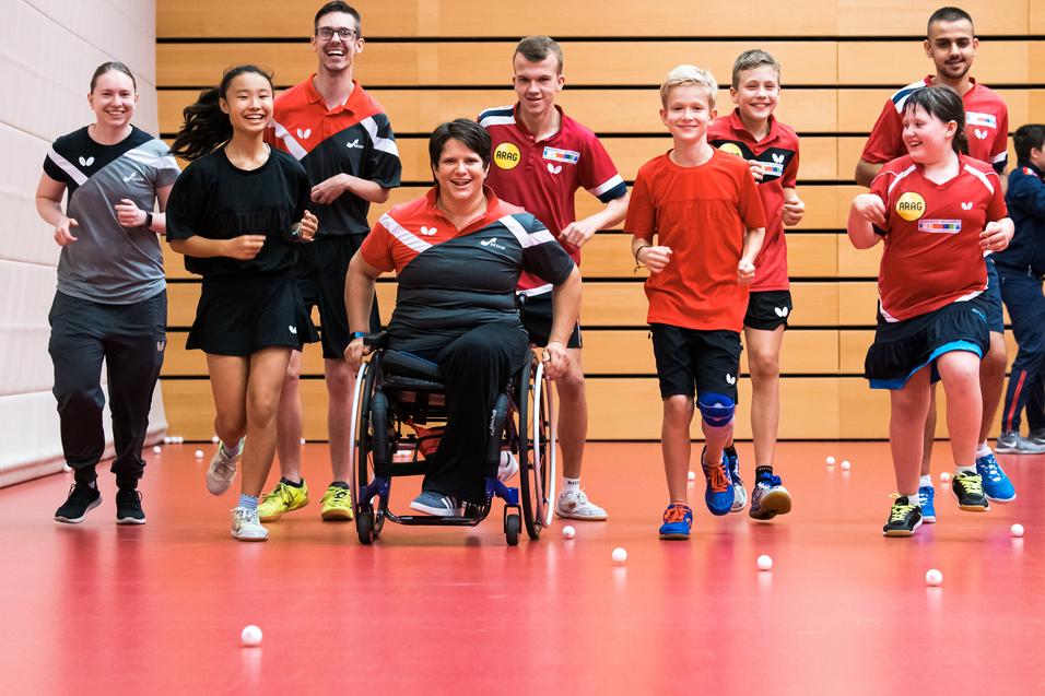 Gruppe von Kindern und Erwachsenen in Sportkleidung joggt in einer Halle mit Tischtennisbällen auf dem Boden.