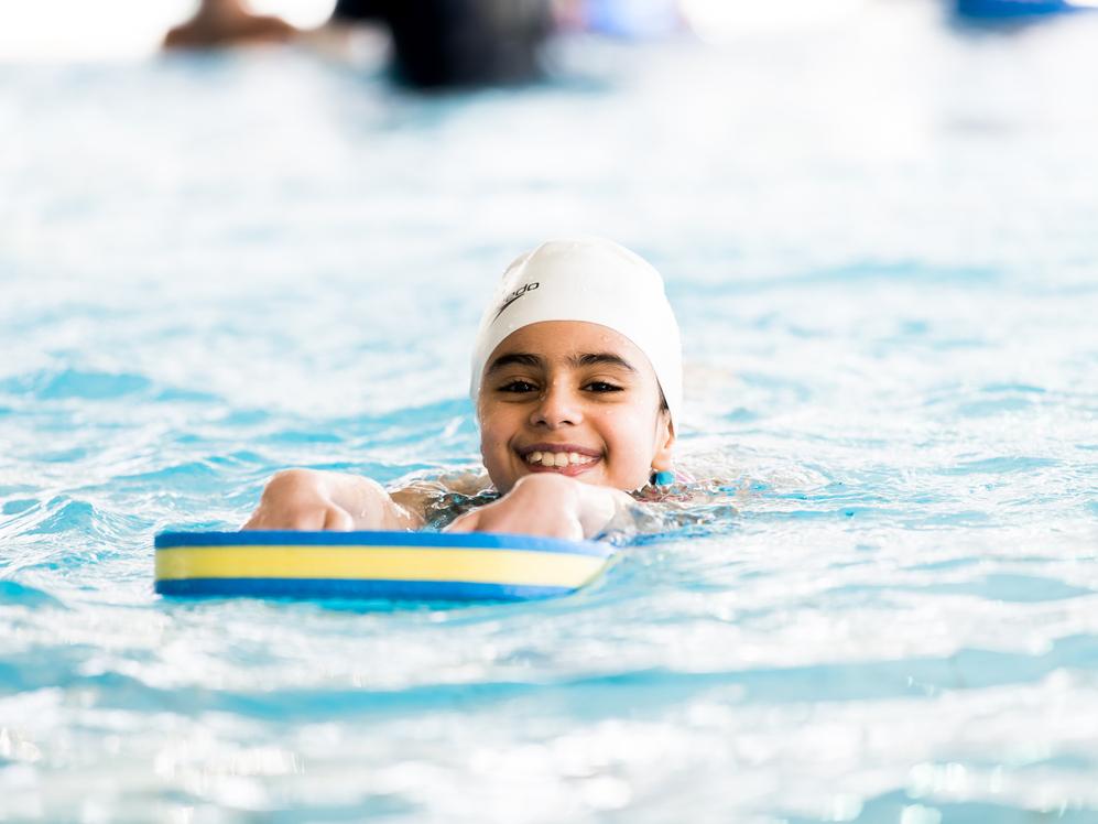 Lächelndes Kind mit Badekappe schwimmt auf einem Schwimmbrett im Wasser eines Hallenbades.